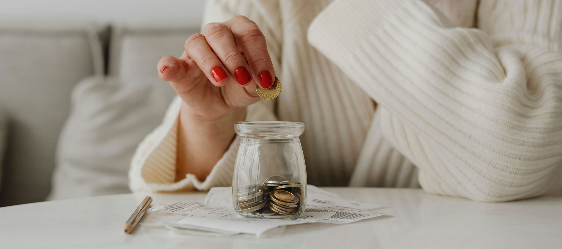 A hand with red nail polish places a coin into a small glass jar filled with coins, resting on scattered papers and a white table in a cozy room.