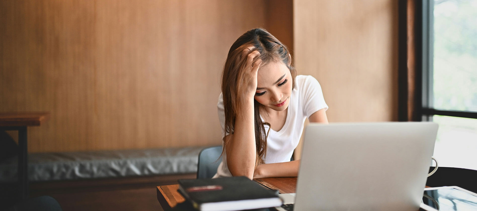 A person sits at a desk, resting their head on one hand while looking at a laptop screen, in a warmly lit room with wooden walls.