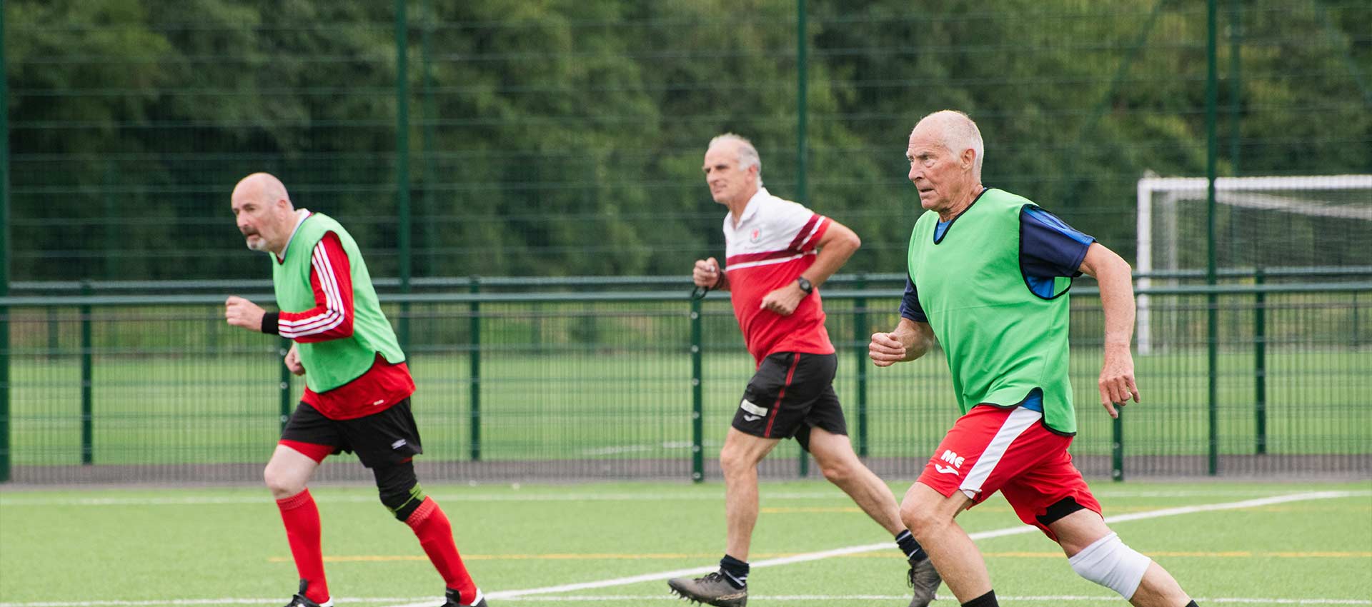 Three older men dressed in red, white and black football kits running across an astroturf football pitch.