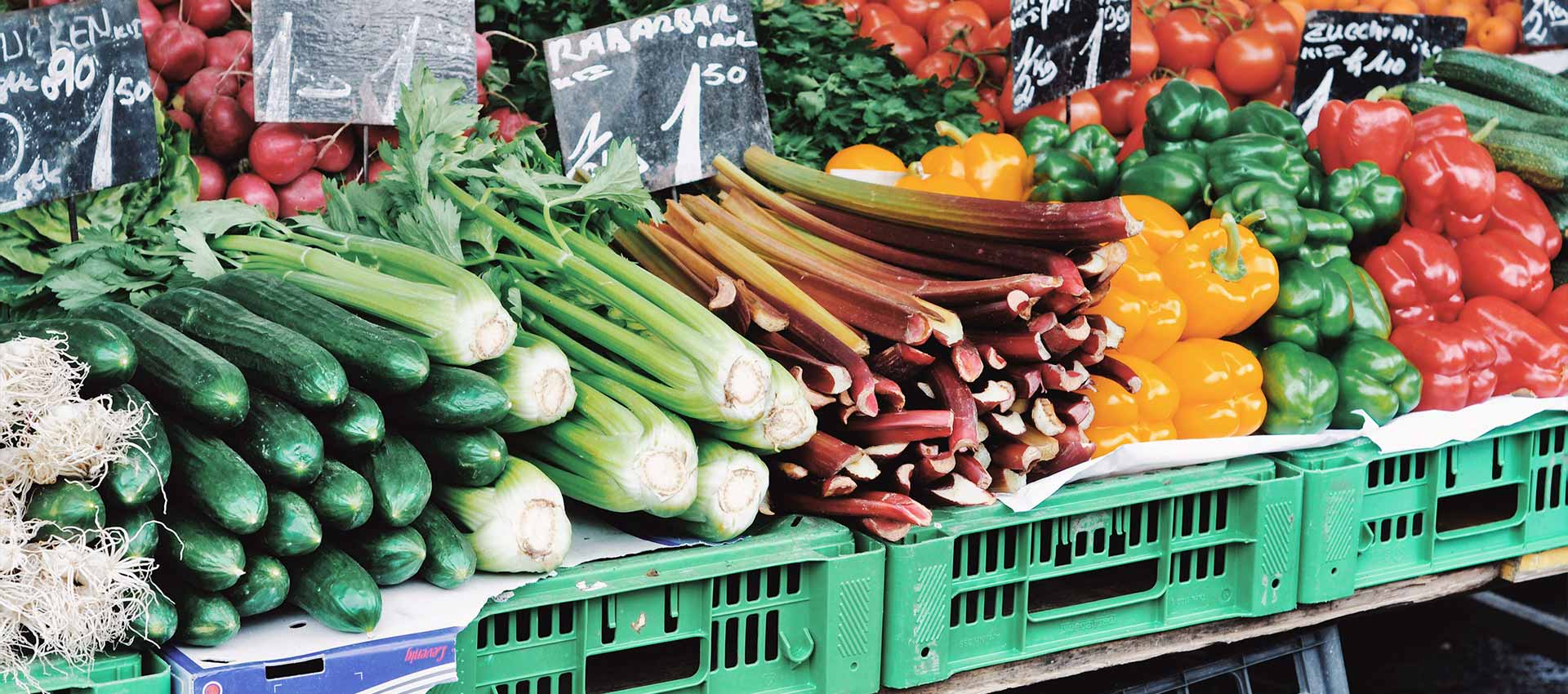 Close up of a vegetable stall with cucumber, celery, rhubarb and orange, green and red peppers stacked in green containers.
