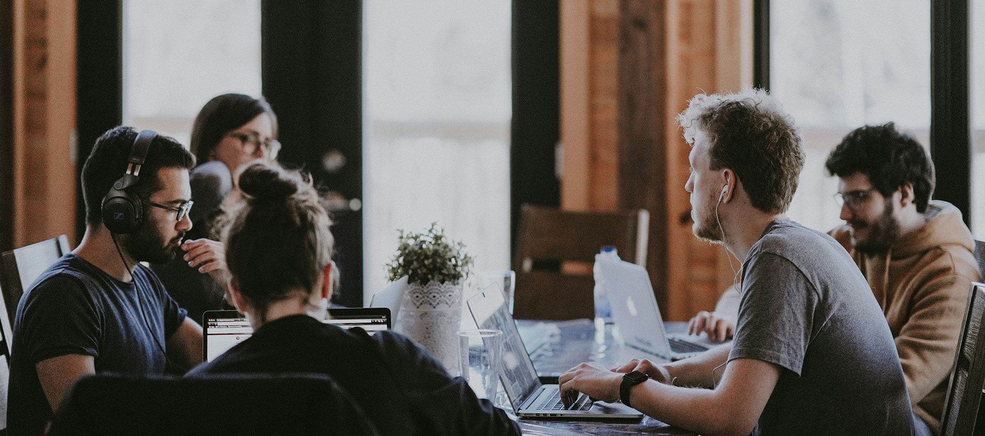 People collaborate around a table with laptops in a cozy office setting. One person is wearing headphones, while others are engaged, reflecting teamwork and productivity.