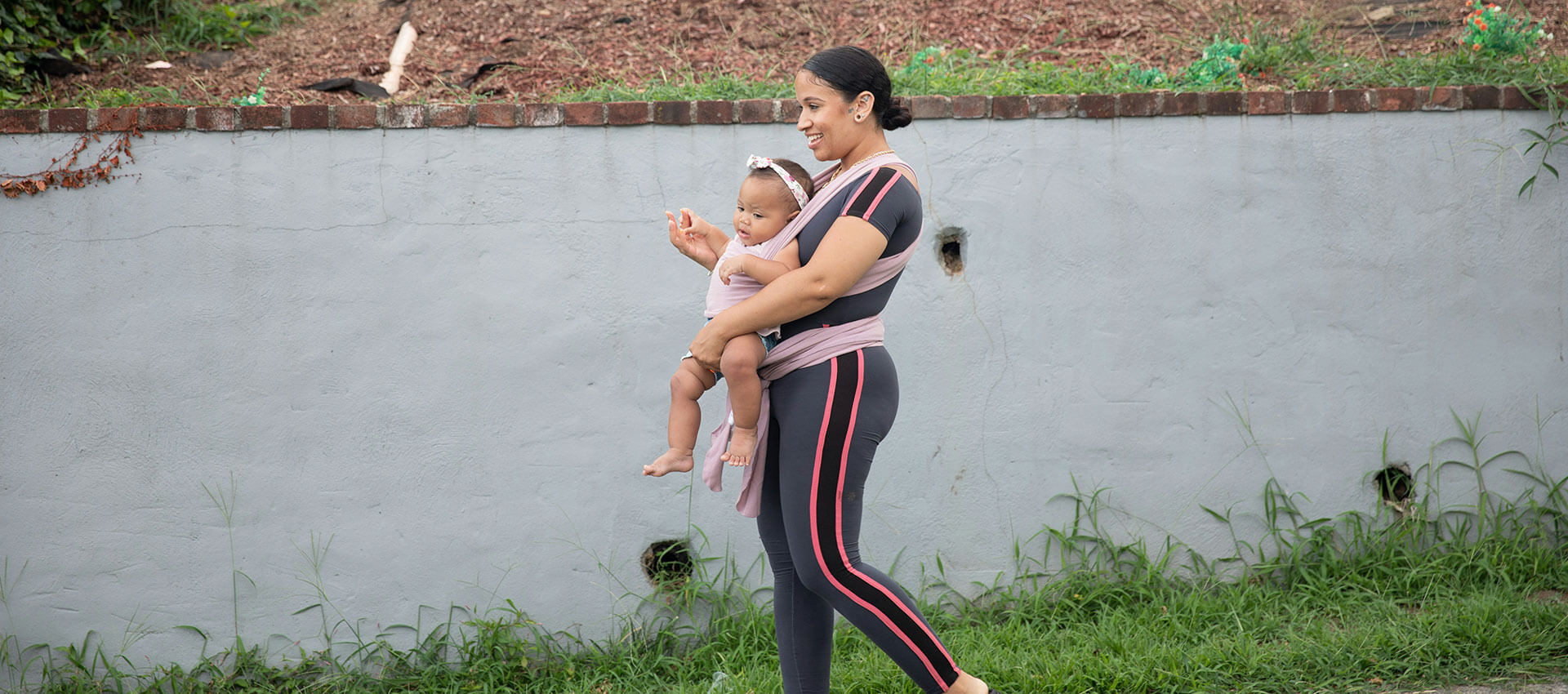 A person walks with a baby in a sling in front of a grey wall, surrounded by grass.