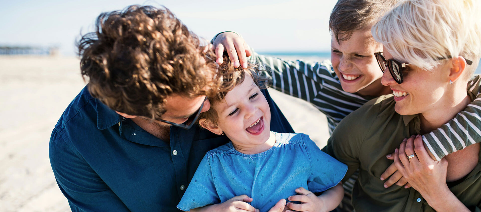 A family of four, including two adults and two children, smiles and embraces playfully on a sunny beach, creating a joyful and relaxed atmosphere.