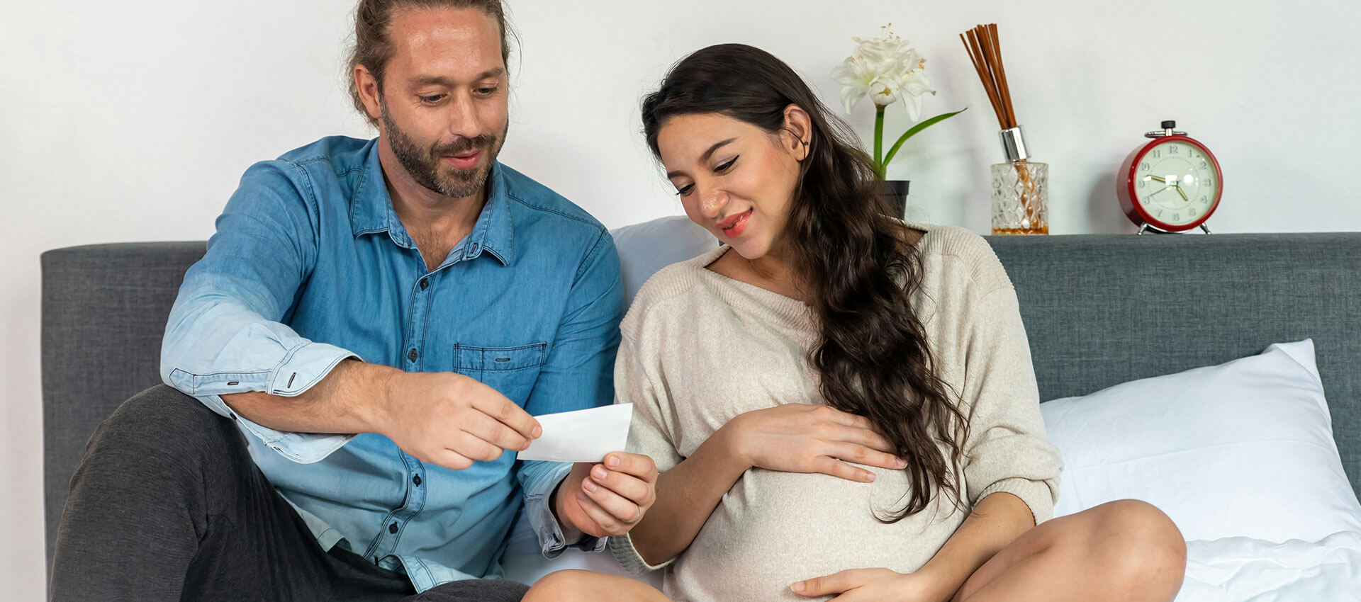 A couple sits on a sofa, with the man showing a photograph to the pregnant woman. In the background, there's a vase with flowers, a decorative object, and a wall clock.