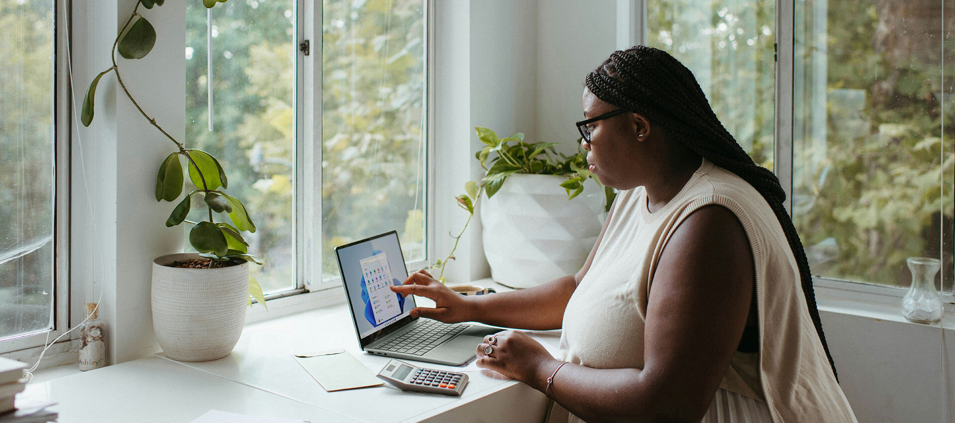 A person sits at a desk by large windows, typing on a laptop. A calculator and potted plants are nearby, with a view of greenery outside.