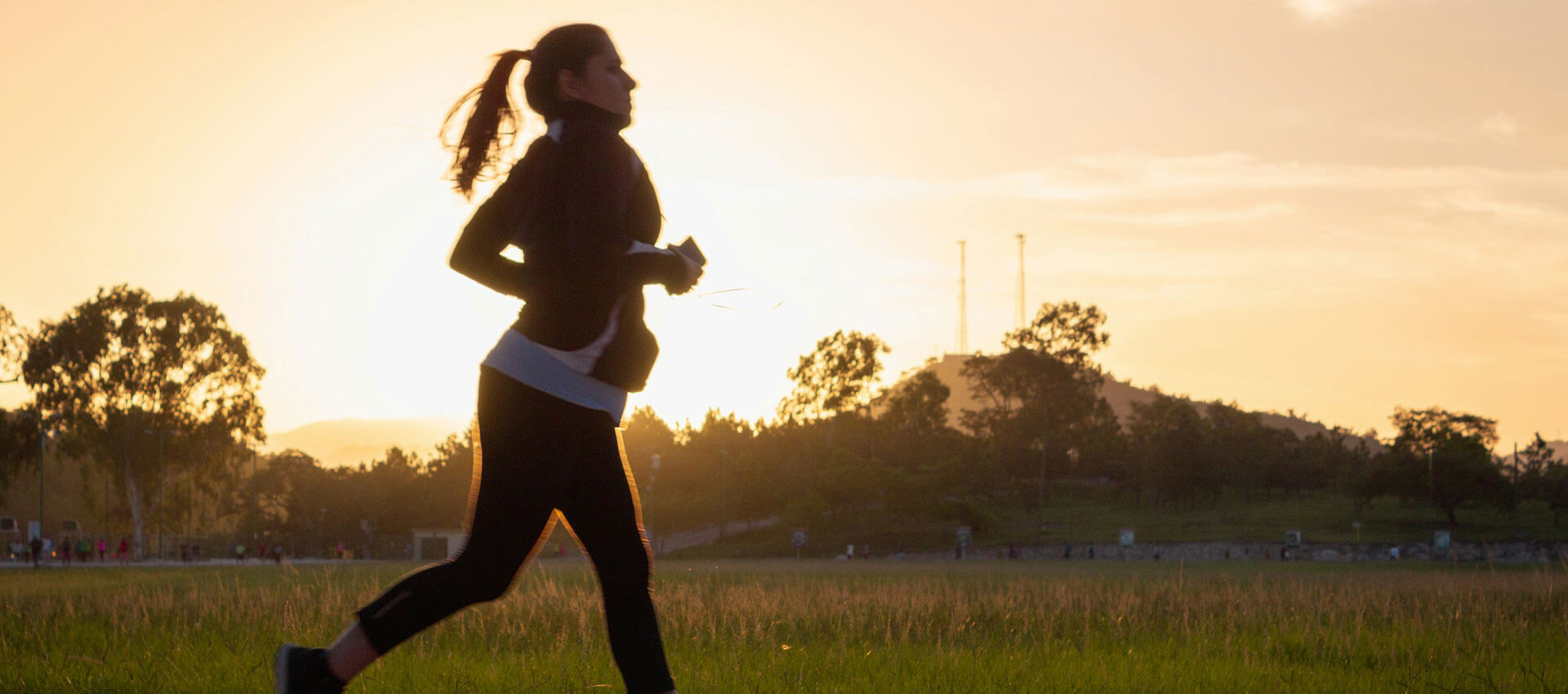 A person jogs in a grassy field at sunset, silhouetted against the glowing sky with trees and a hill in the background.