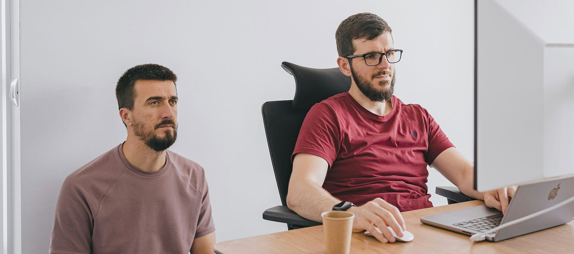 Two men focus intently on a large computer screen; one sits at a desk chair while the other kneels beside him in a white-walled office setting.
