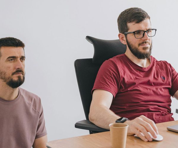 Two men focus intently on a large computer screen; one sits at a desk chair while the other kneels beside him in a white-walled office setting.