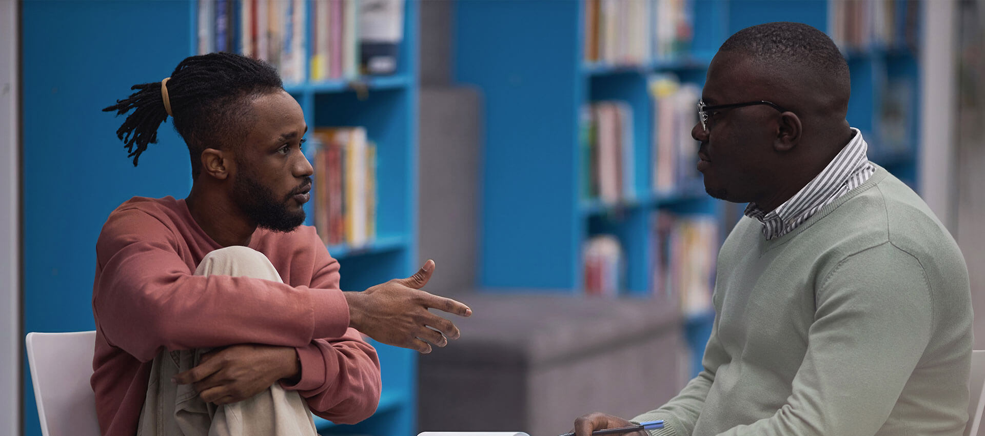 A person in a red jacket and white mask sitting on a chair raises a hand, while another person in an orange mask listens attentively. They are in a room with blue walls.