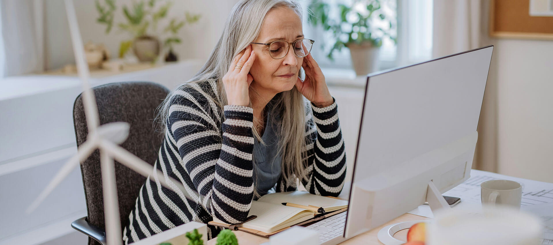 A person with long hair and glasses sits at a desk, massaging their temples while looking at a computer monitor. The room is bright with plants and papers nearby.