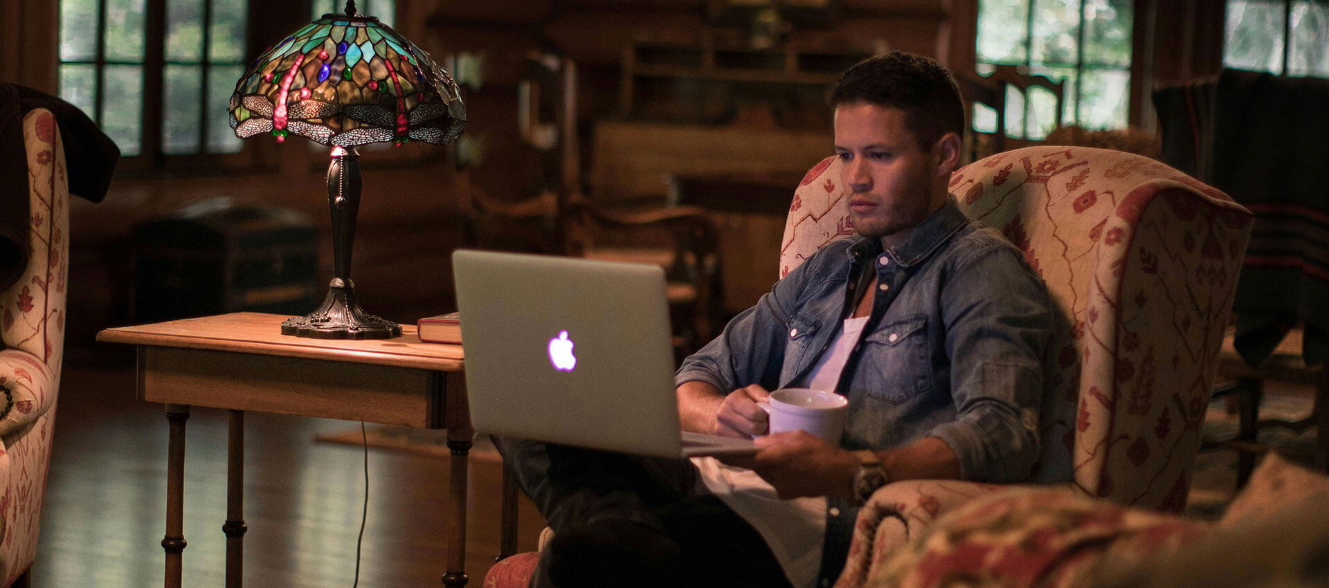 A man sits in an armchair, using a laptop, holding a mug. A decorative table lamp glows beside him, within a cozy room featuring wooden furniture and large windows.
