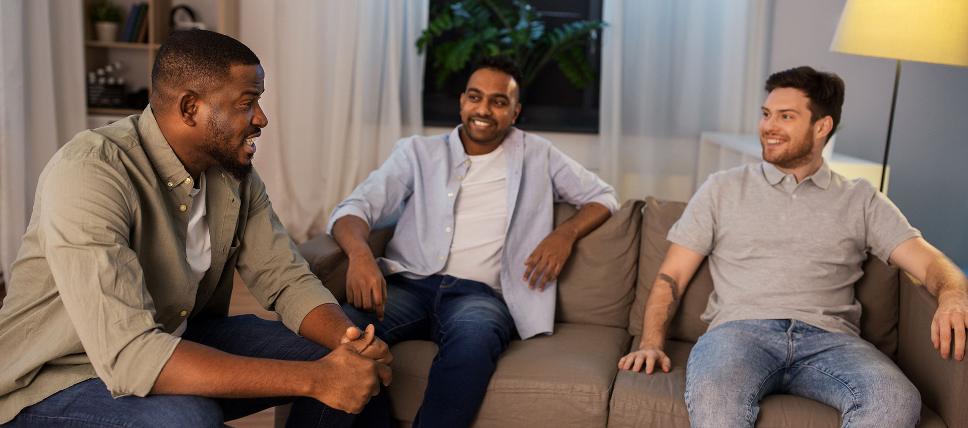 Three men sat in a living room on sofas chatting and smiling.