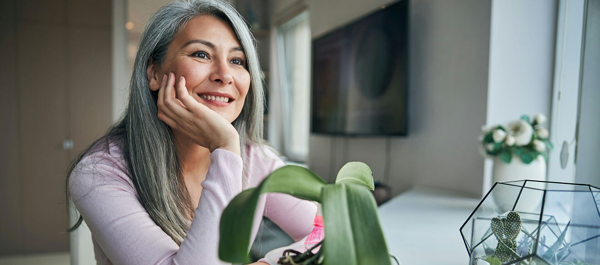 A person with long blonde hair is smiling warmly, standing against a textured, light brown wall. They are wearing a light-colored shirt with subtle patterns.