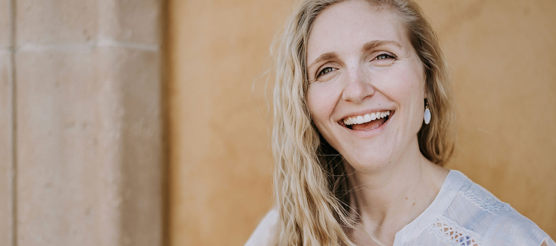 A person is smiling warmly, standing against a light brown wall. Their expression conveys positivity and friendliness, creating a welcoming atmosphere.
