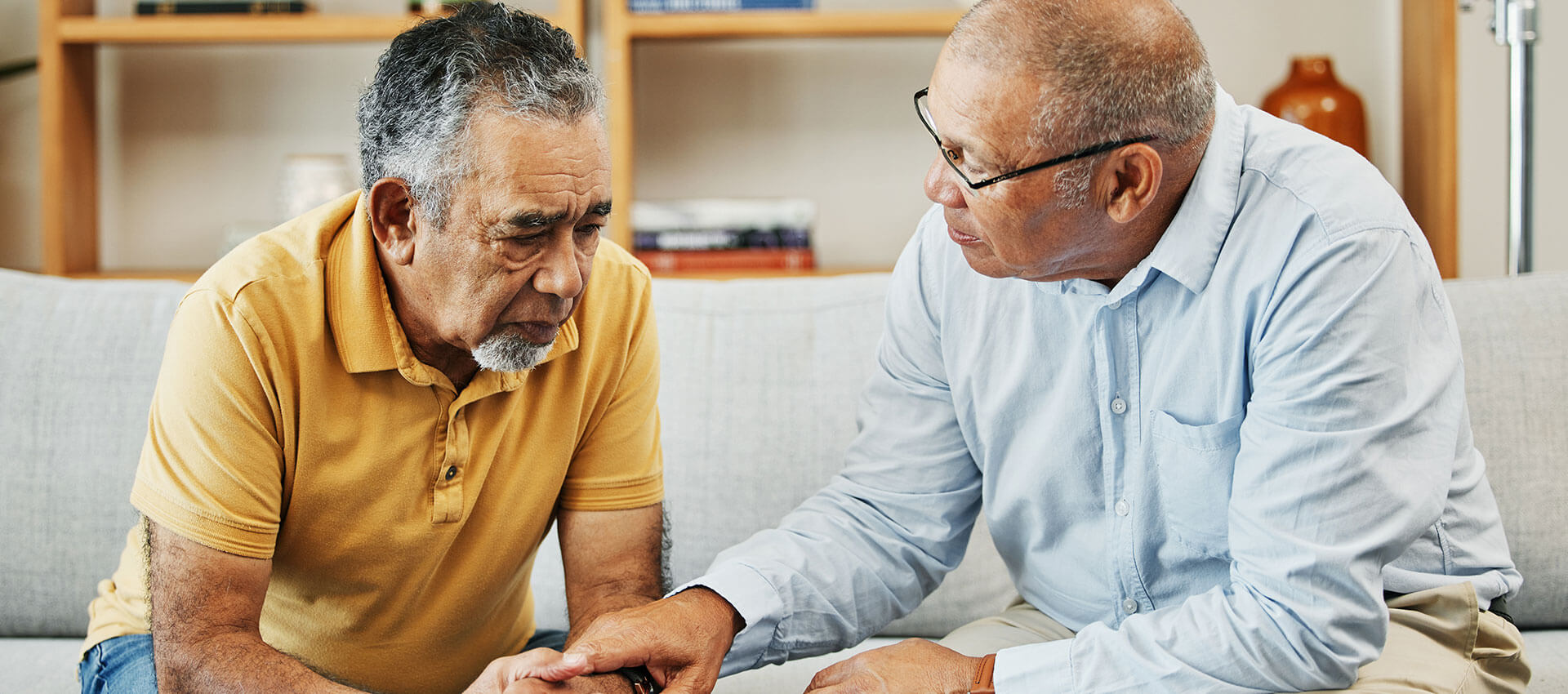 Two elderly men sit closely on a sofa, one wearing a yellow shirt, appearing concerned, while the other in a blue shirt, offers comfort by holding his hand. Shelves with books are behind them.
