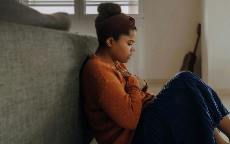 A person sits on the floor, leaning against a sofa with their knees drawn up, in a softly lit room with a guitar and window shutters in the background.