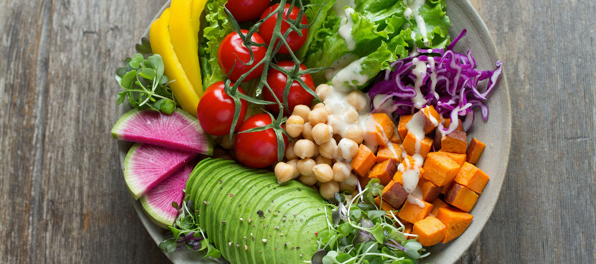 A bowl contains sliced avocado, radishes, cherry tomatoes, yellow bell pepper, chickpeas, cubed sweet potatoes, and lettuce. Drizzled with dressing, it rests on a rustic wooden surface.