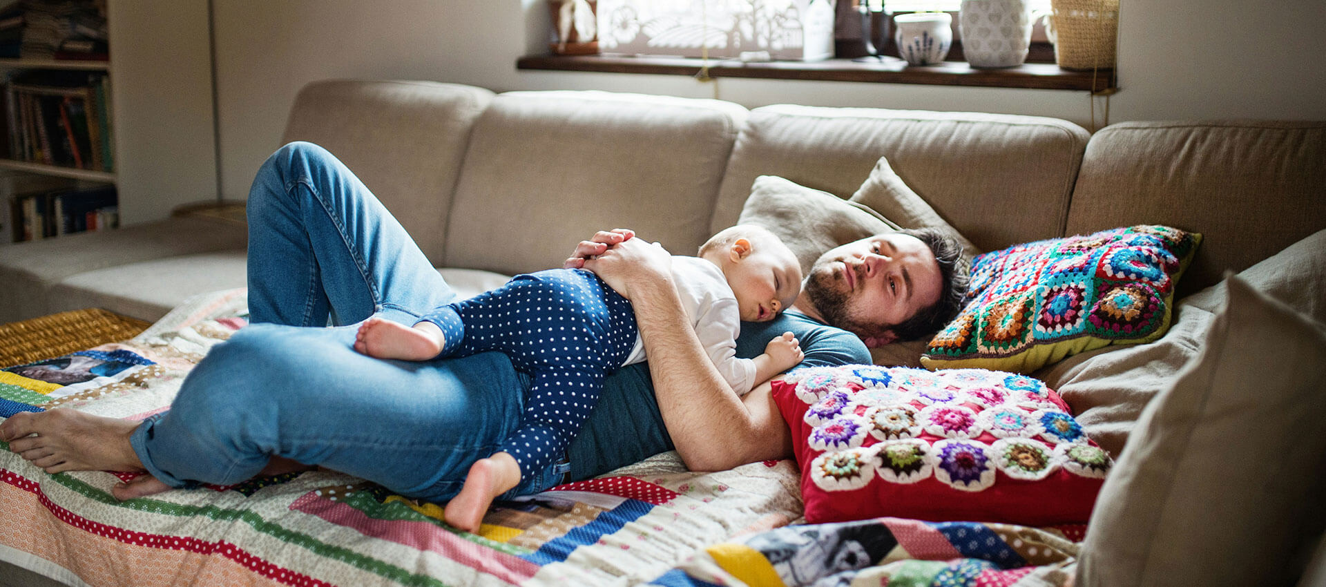 A bearded man lies on a patterned sofa, gently holding a sleeping baby on his chest. Colorful crochet cushions surround them in a cozy living room setting.