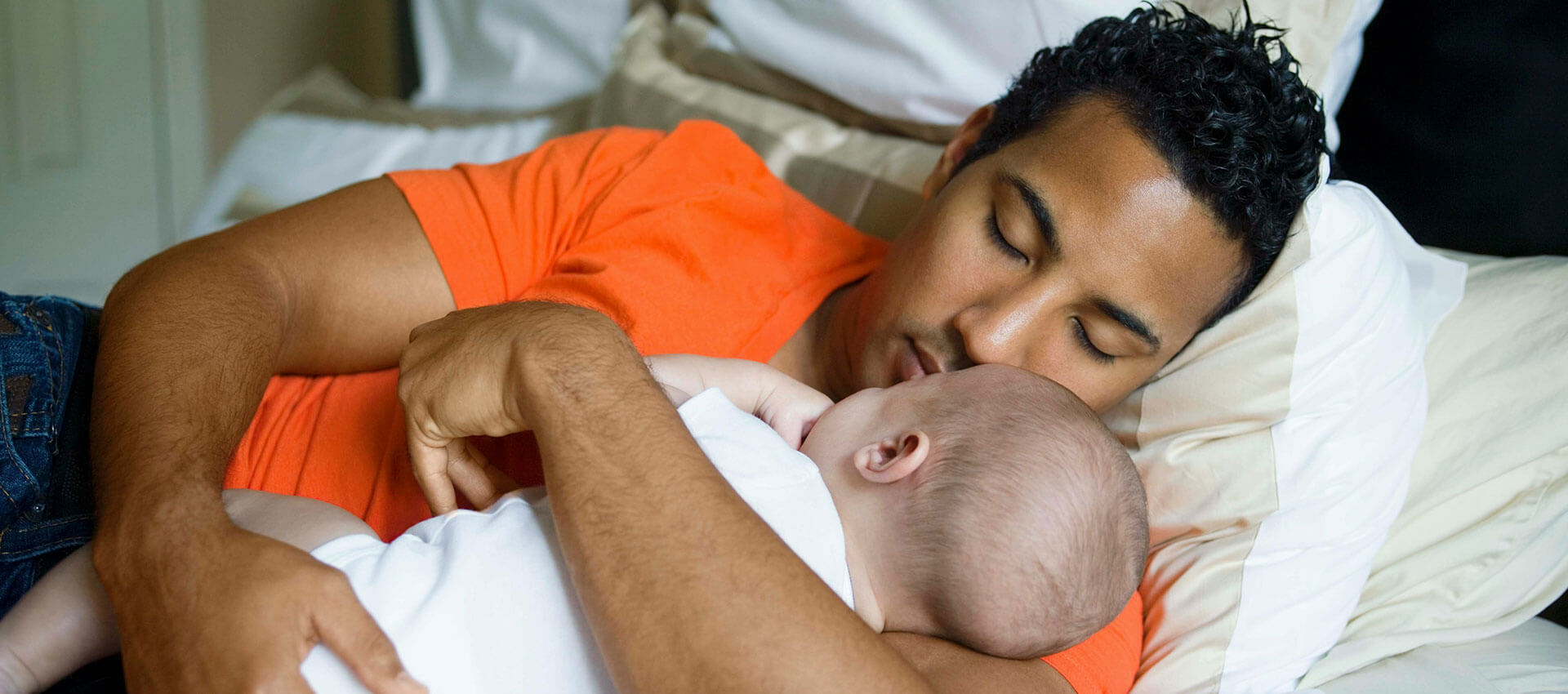 A man in an orange shirt is lying on a bed, sleeping while cradling a baby in his arms. They are surrounded by white and beige pillows.