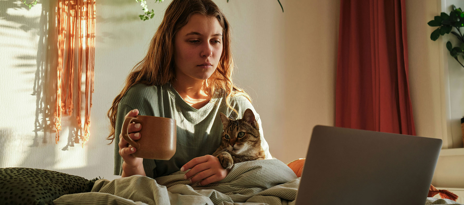 A young woman sit in her bed with a cup in her hand, a cat on her lap and a laptop in front of her.