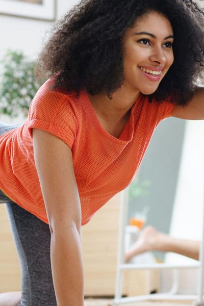 Person in a bright orange top performs a yoga pose, extending an arm forward, in a bright room with plants and wooden elements in the background.