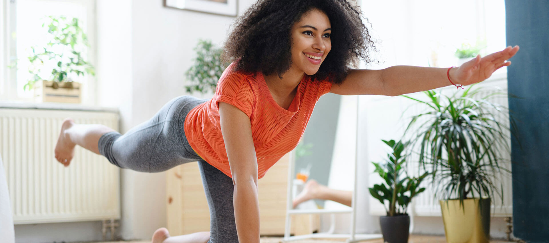 Person in a bright orange top performs a yoga pose, extending an arm forward, in a bright room with plants and wooden elements in the background.