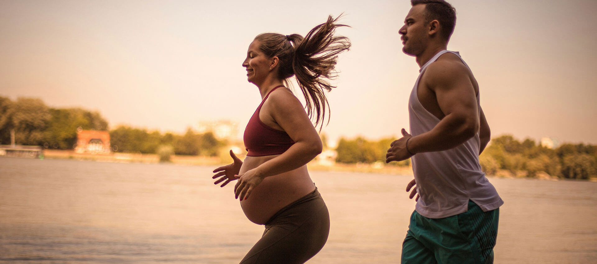 A pregnant woman and a man are jogging along a lakeside path, surrounded by greenery and a calm body of water under a warm sky.