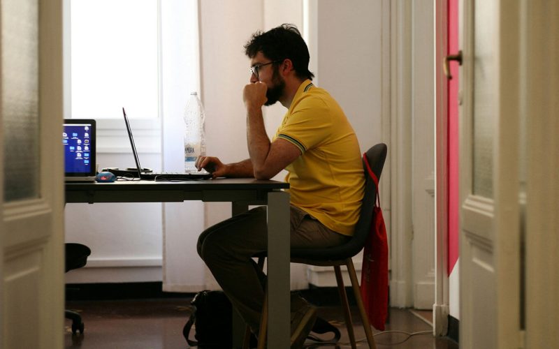 A person in a yellow shirt sits at a desk, using a laptop, in a well-lit room with white walls and a visible bottle of water.