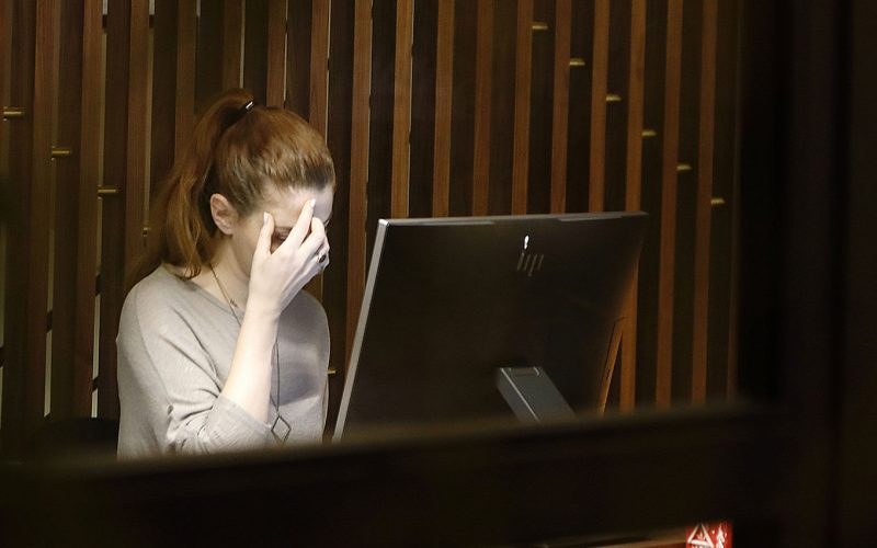 A person sits at a computer with their hand on their face, looking stressed. They are in a dimly lit room with wooden paneling. No text is visible.