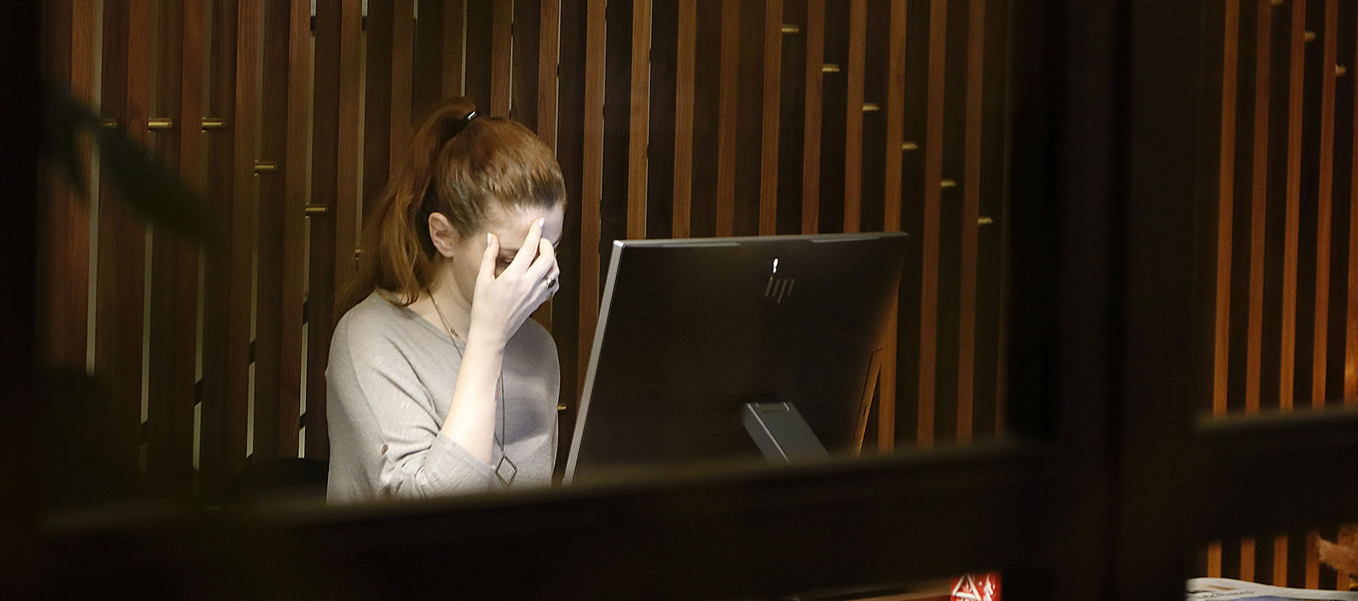 A person sits at a computer with their hand on their face, looking stressed. They are in a dimly lit room with wooden paneling. No text is visible.