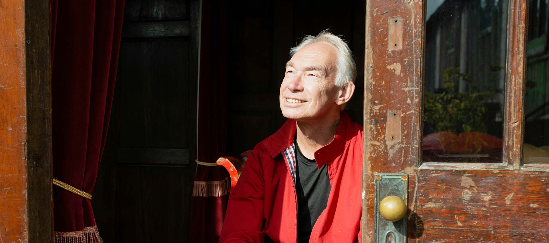 An older grey haired man in a red jacket sat on chair in the doorway of an old building looking up in to the daylight.