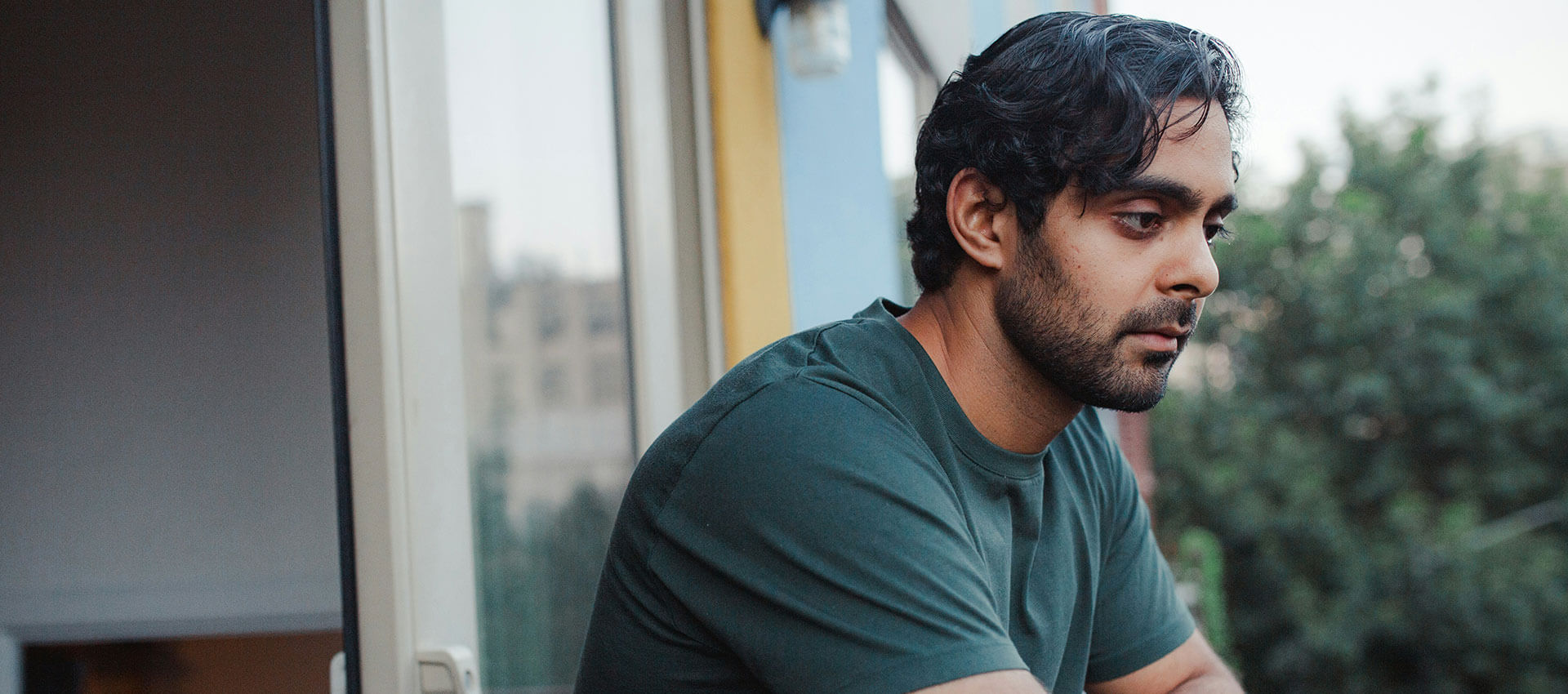 A man in a green shirt gazes pensively from a balcony, surrounded by trees and buildings in the distance.