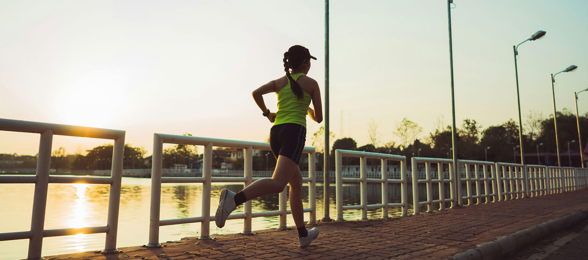 A person runs along a paved path beside a river at sunrise, surrounded by railing and streetlights.
