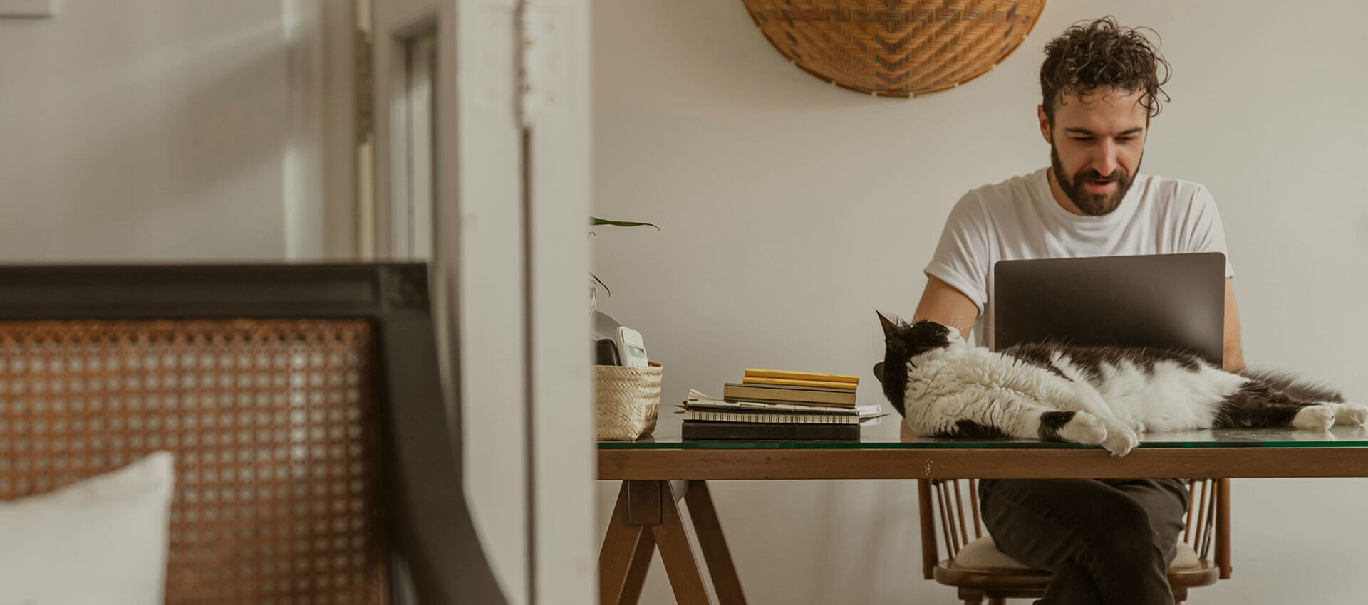 A man types on a laptop at a wooden table, accompanied by a black and white cat lying beside several notebooks and a small basket in a cozy room.