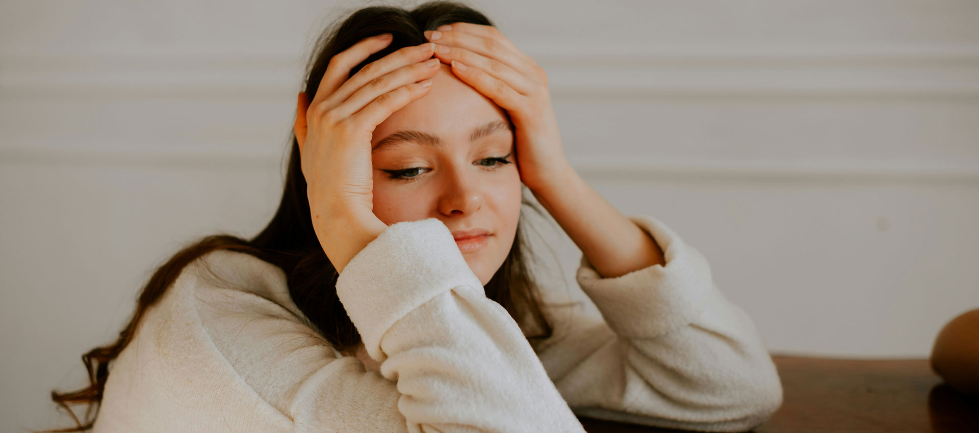 A young woman with dark brown hair has her head in her hands as she sits at a wooden table.