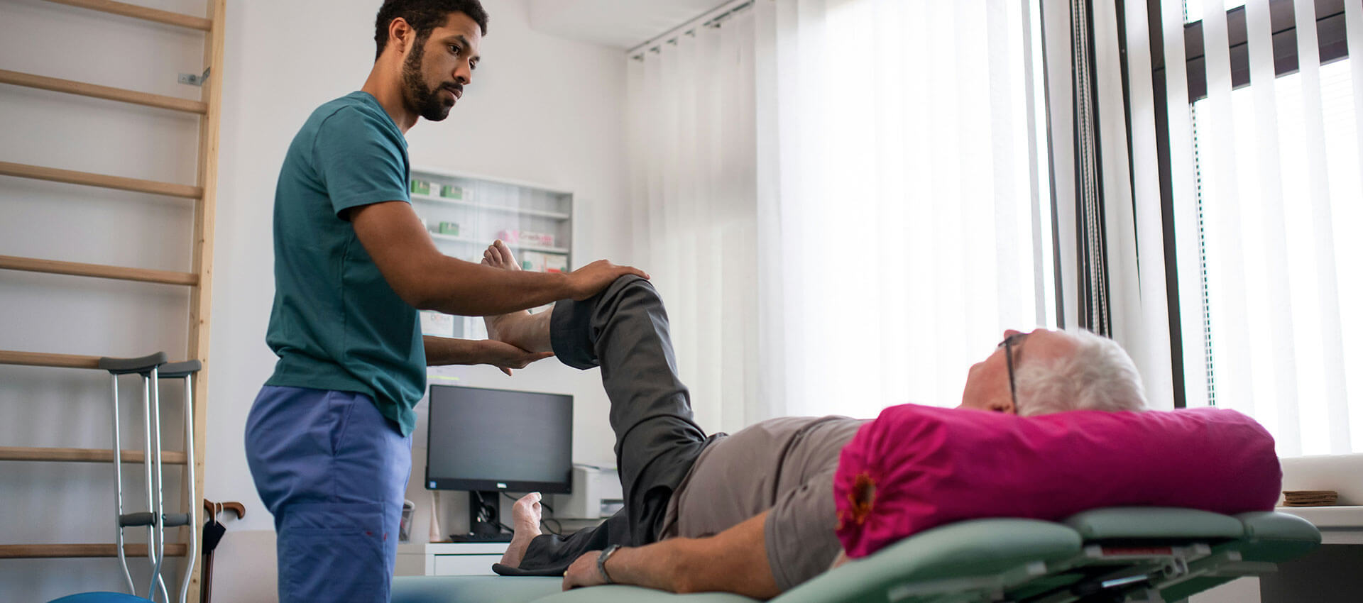 A healthcare professional assists an elderly patient with a leg exercise in a bright, well-equipped therapy room with medical equipment and a computer in the background.