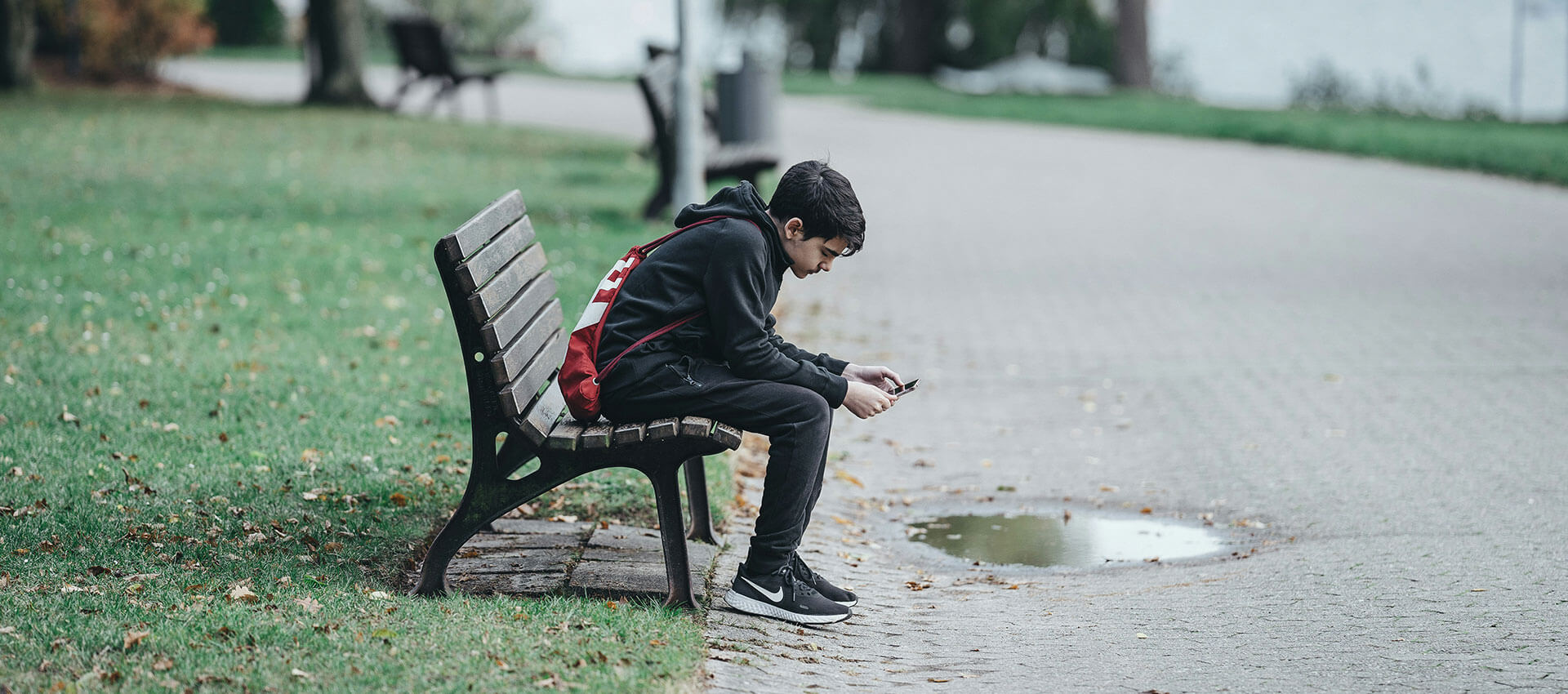 A young person in a black outfit sits on a park bench, looking at a smartphone. A red backpack is slung over their back, and autumn leaves are scattered on the pathway.