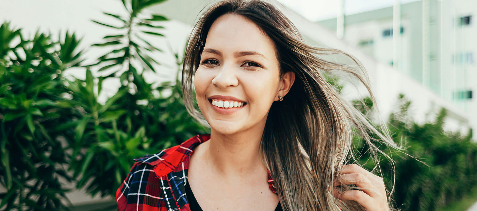 A person smiles outdoors, hand touching their hair, with greenery and blurred buildings in the background.