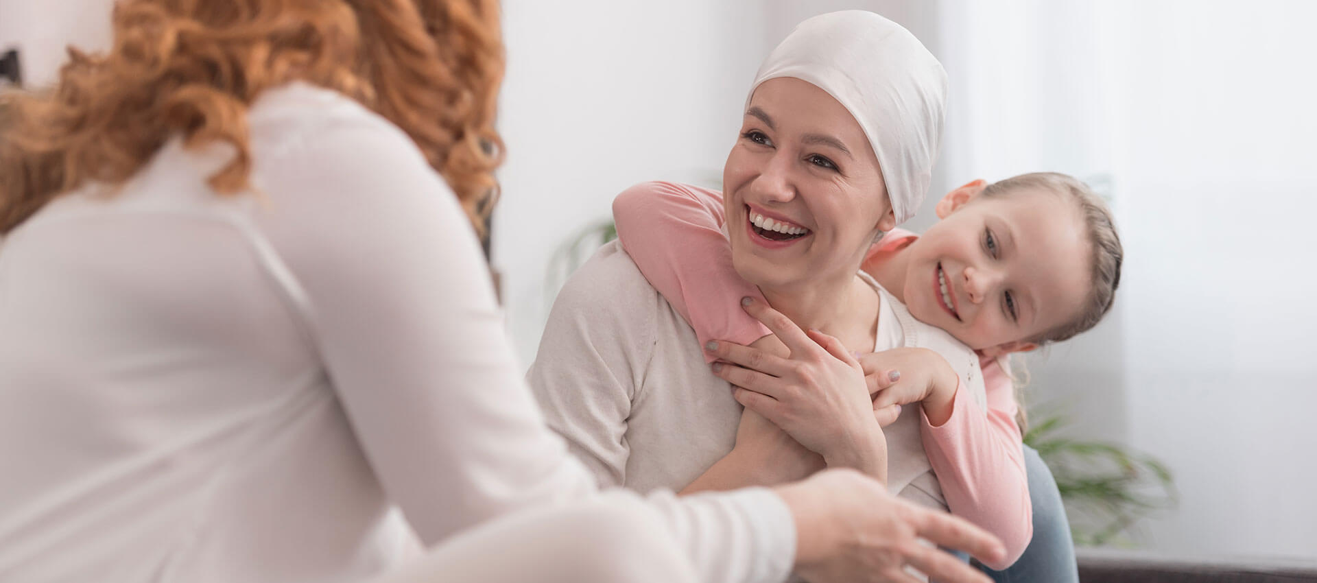 Woman wearing headscarf smiles with a child hugging her from behind, while sitting indoors. Another person with long hair gestures while facing them, creating a warm, supportive atmosphere.