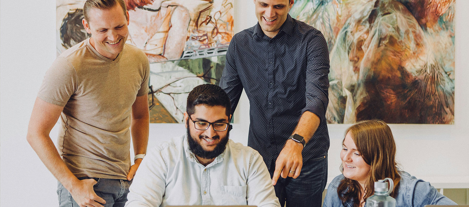 Four people smiling and working around a laptop in an office with abstract art on the wall. A water bottle is on the table, signifying a collaborative meeting.