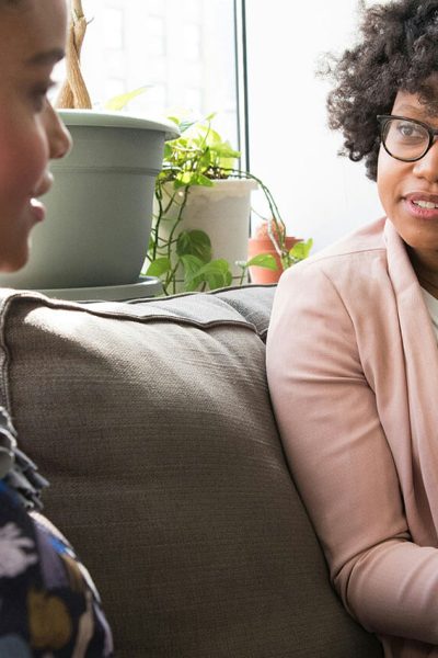 Two individuals are seated on a couch engaged in conversation. One is speaking while the other listens attentively. The setting includes potted plants and a bright window in the background.