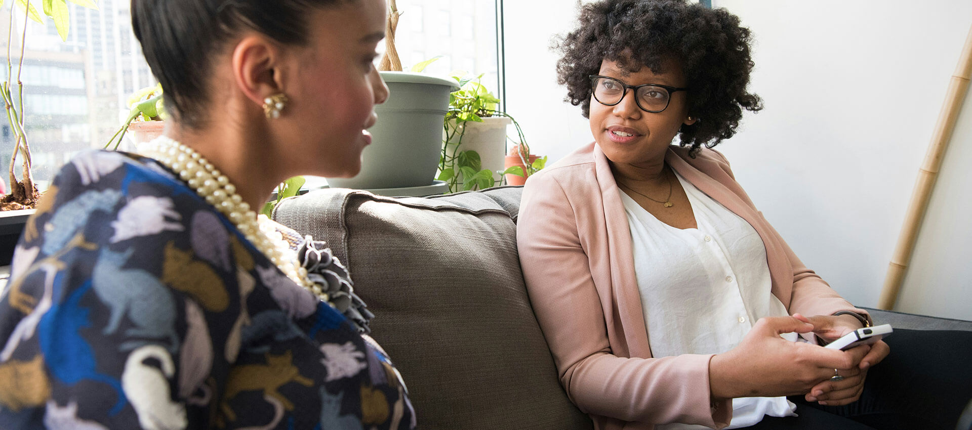 Two individuals are seated on a couch engaged in conversation. One is speaking while the other listens attentively. The setting includes potted plants and a bright window in the background.