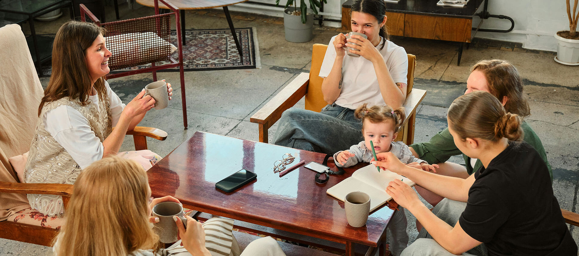 A child and three adults sit around a wooden coffee table, sipping drinks and reading in a cozy, sunlit living room with a patterned rug and potted plant.