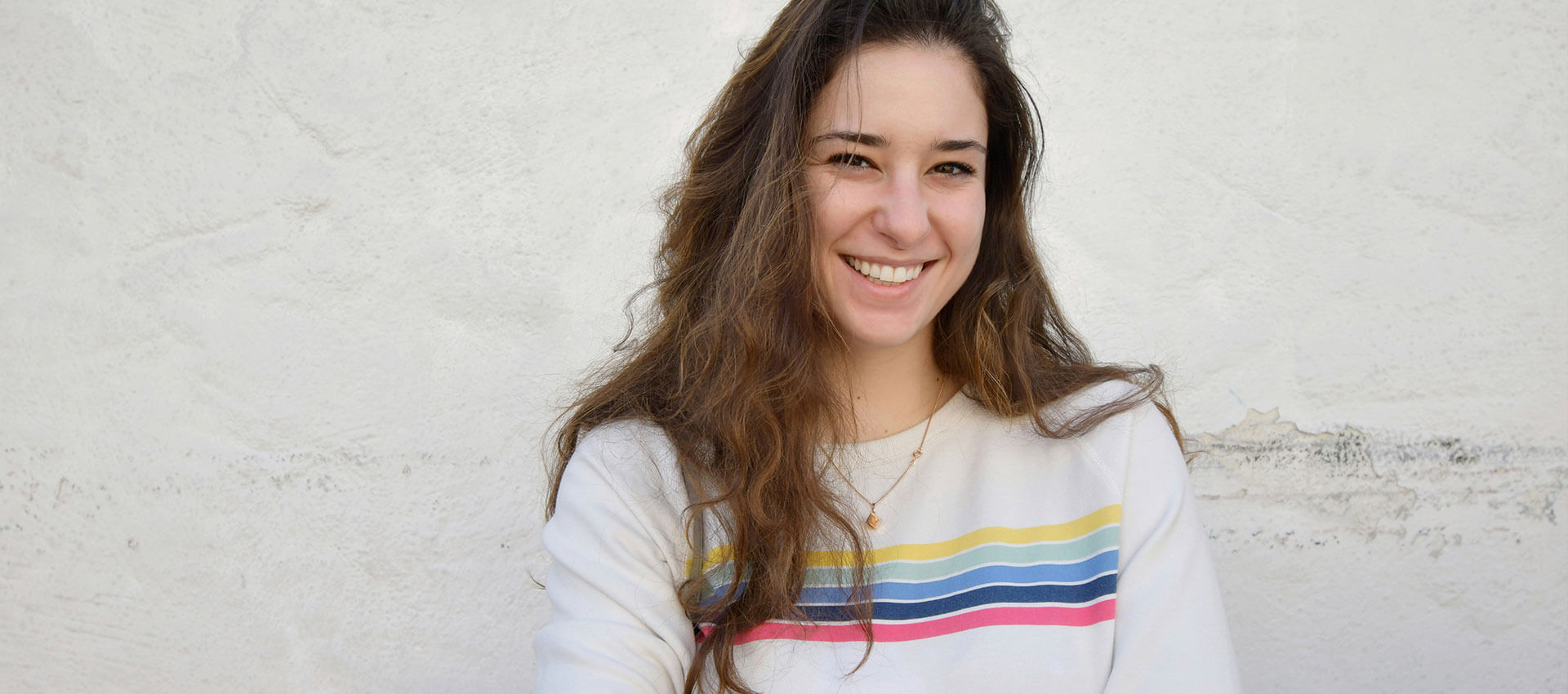 A woman with long brown hair smiles warmly, wearing a white sweater with colorful stripes. She stands against a textured white wall background.