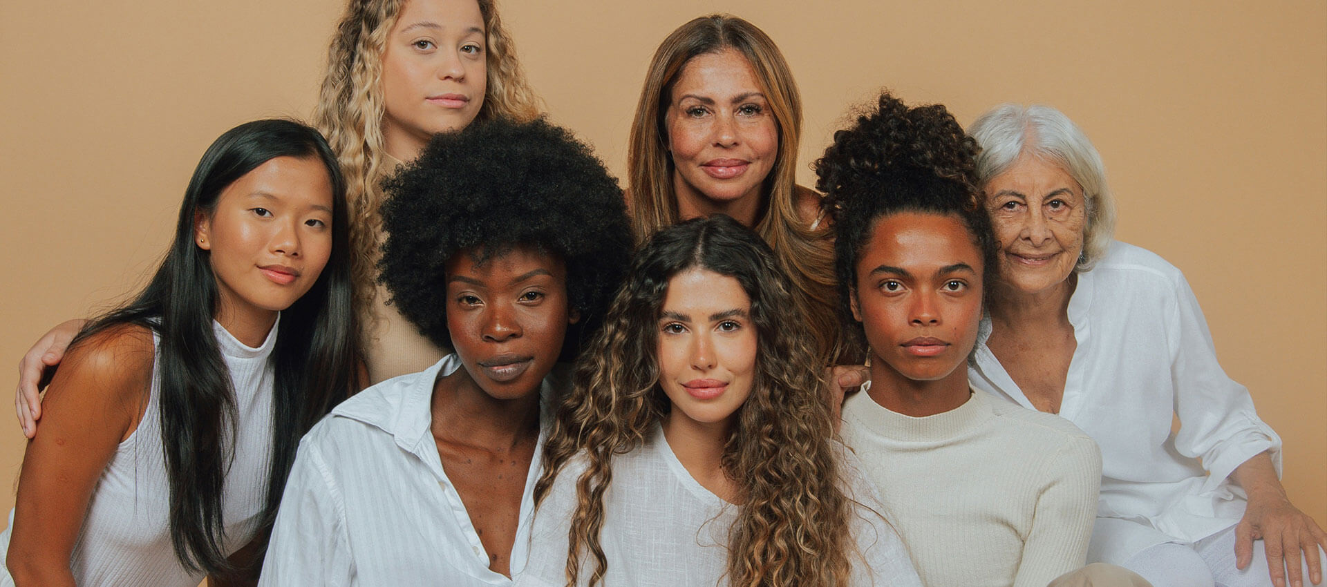 A diverse group of seven women of various ages and backgrounds are sitting closely together, smiling gently for the camera, against a plain beige background. They wear casual, light-colored clothing.