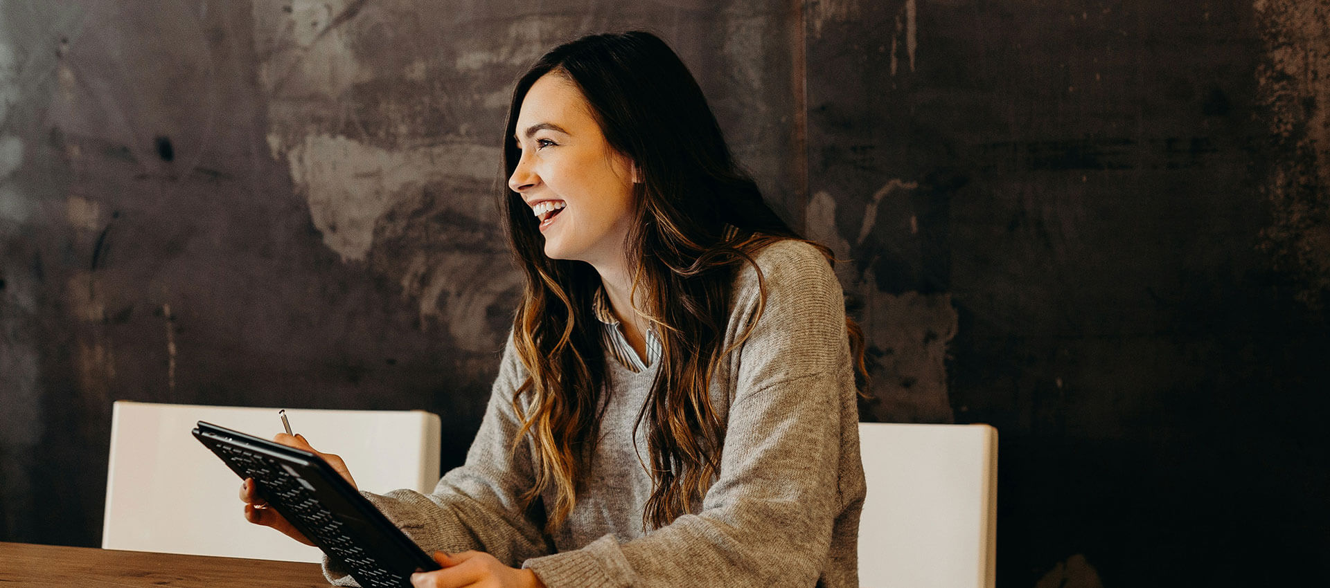 A woman holding a pen and tablet smiles while sitting at a table, against a dark, textured wall.