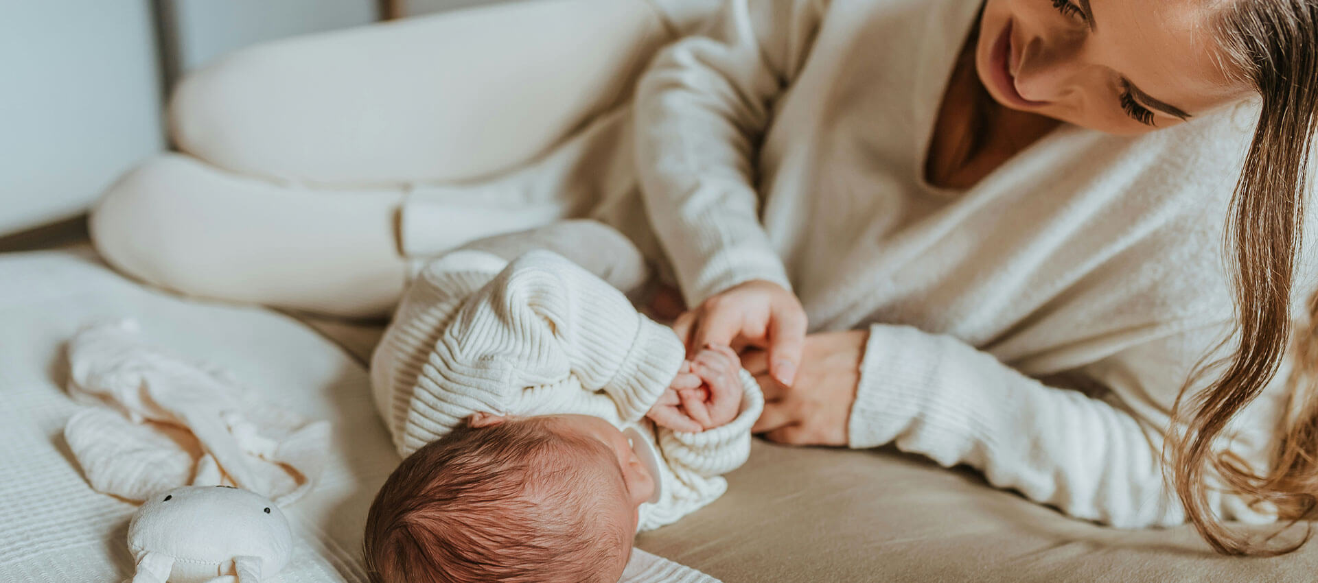 A baby lies on a bed, reaching towards a woman who smiles down at them. The setting is cozy with soft lighting and neutral-colored bedding.