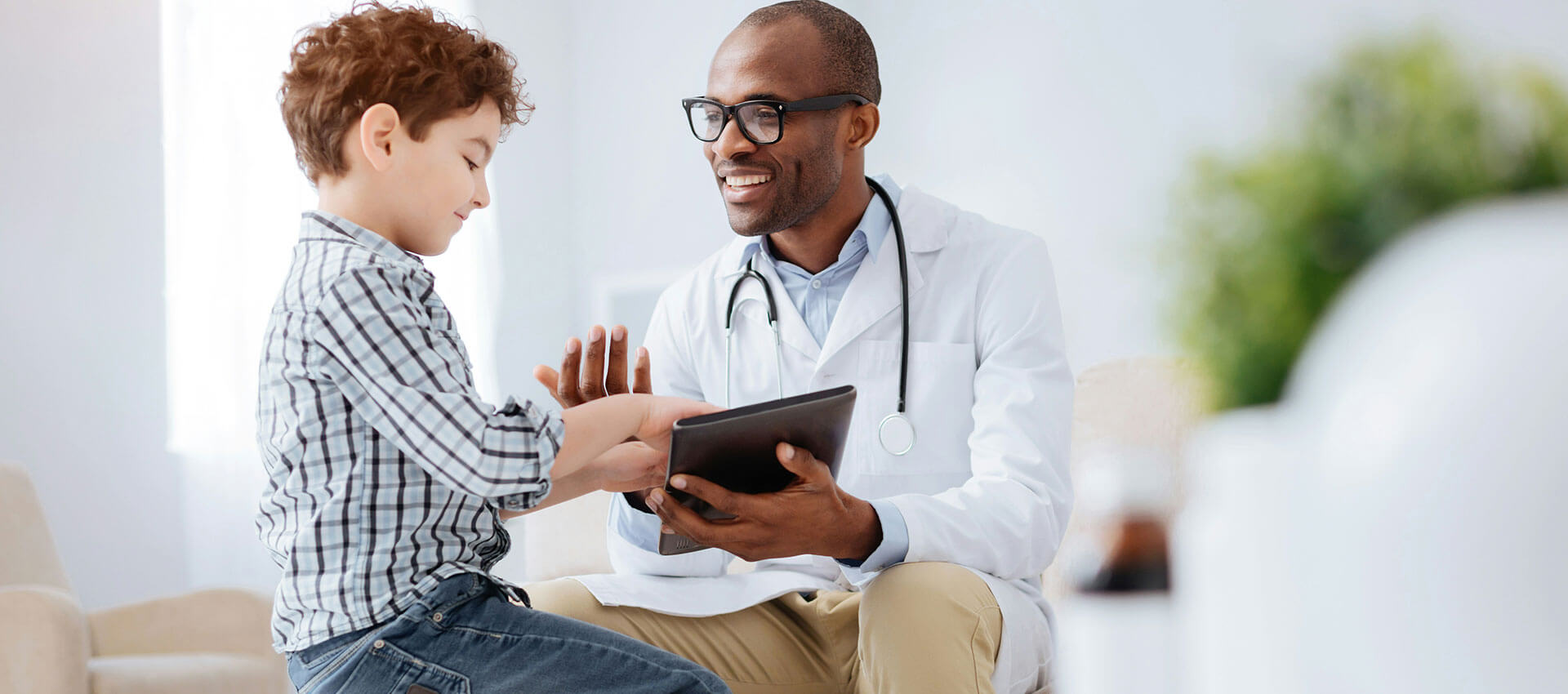 A doctor holds a tablet while talking to a seated child in a plaid shirt, inside a bright medical office.
