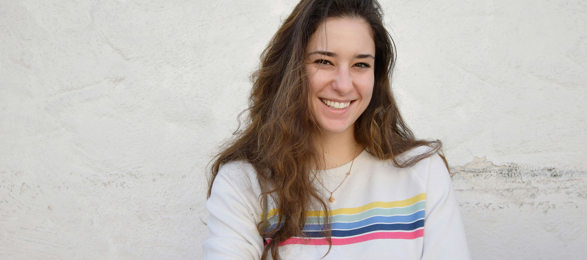 A woman with long brown hair smiles warmly, wearing a white sweater with colorful stripes. She stands against a textured white wall background.