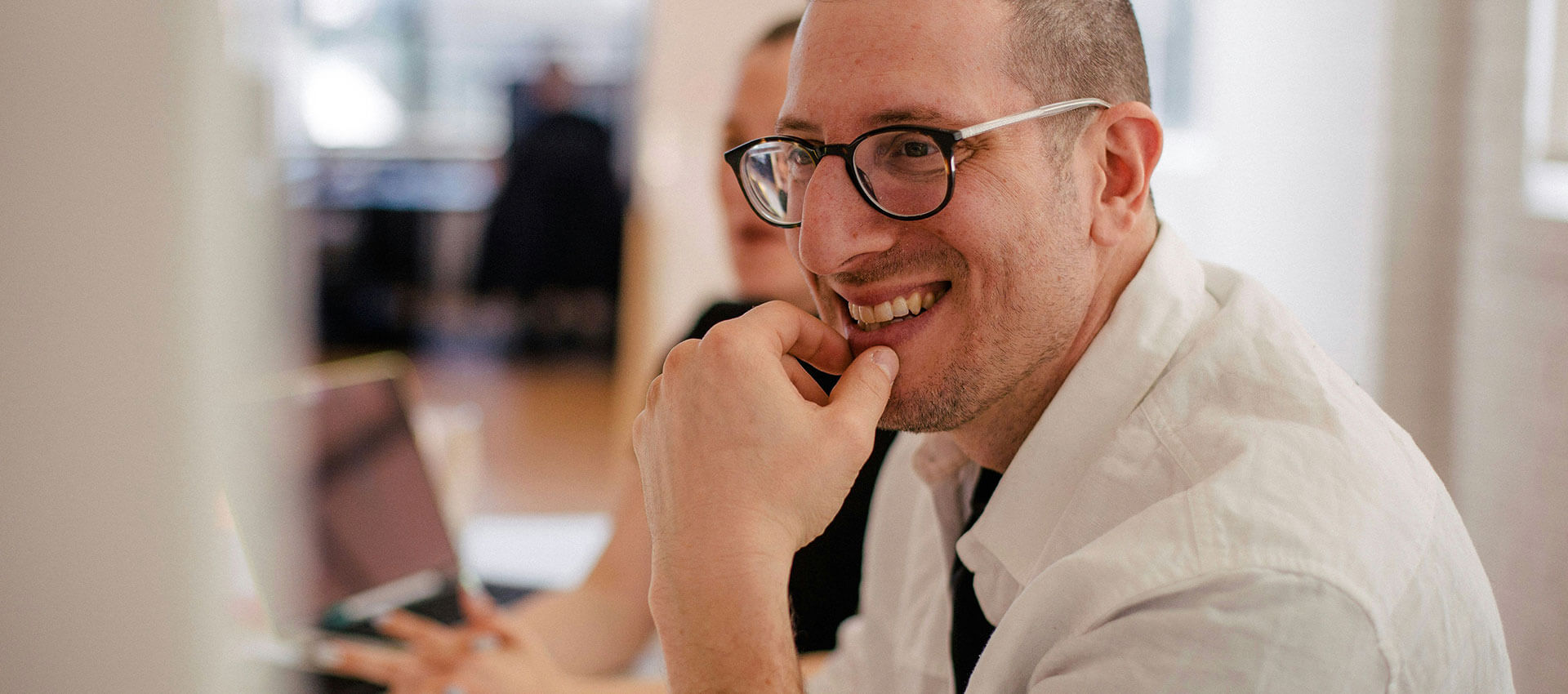 A man wearing glasses smiles with his hand on his chin. He is sitting at a desk with a laptop, in an office environment.
